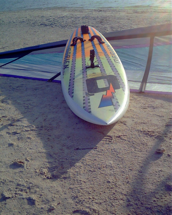 Mike Kimbro's Aerotech 7.5 sail under his vintage 1989 O'Brien Epoxy Lite 9.9 giant slolam windsurfer, as the sun drops low at Corpus Christi's Bird Island Basin sailing and fishing area.
